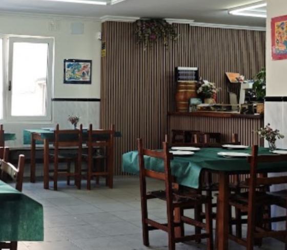 Otra vista del comedor: pared de listones de madera, una barra pequeña y mesas con mantel verde.